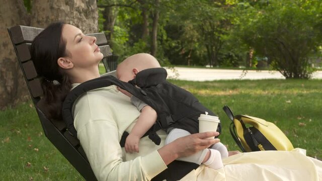 Mother with sleeping baby in infant kid carrier sling resting outdoors in city park, six month old baby boy in baby hipseat. High quality 4k footage