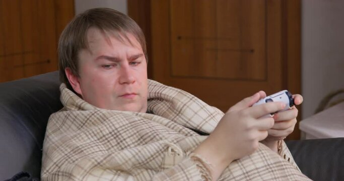 Mature plump man wrapped with warm blanket gets distracted BY dachshund dogs and loses game sitting in armchair with wireless controller