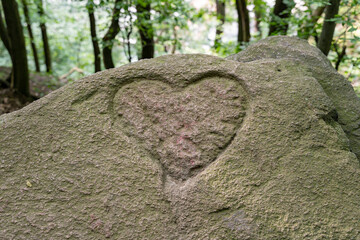 A heart is engraved in a big natural stone. The sign for love in a huge rock in the middle of a forest. Trees and green leaves in the background. A carved symbol in a block.