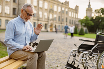 Senior man, recovering patient with wheelchair having video call, working on a laptop while sitting on the bench in the park near clinic