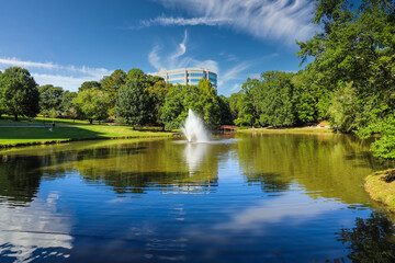 Naklejka na ściany i meble a breathtaking shot of a lake with a water fountain in the center of the lake with lush green and autumn colored trees reflecting off the water with blue sky and clouds at Lenox Park in Georgia