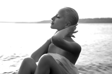 Art nude sexy blonde with short haircut is sitting in water on shore beach of lake at sunset. Wet hair and a woman body. Secluded beach holiday. Black and white - Powered by Adobe