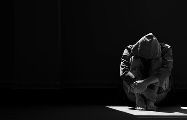 Light and shadow on surface of hopeless man in hoody sitting alone with hugging his knees on the floor in empty dark room in black and white style - obrazy, fototapety, plakaty
