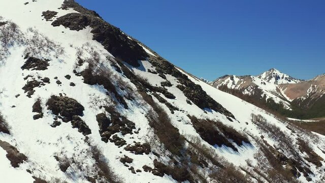 Aerial drone dolly shot descending down the side of a mountain side in melt in spring at the Cerro Bayo ski resort near Villa La Angostura, revealing forest and rocks on the mountains