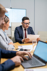 Serious mature broker looking through financial papers in front of young employees