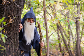 A wizard with a long gray beard and a cloak in a deep forest. An elderly man in a witcher costume