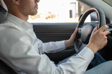 Hands of young elegant man in white shirt holding by steering wheel while driving car