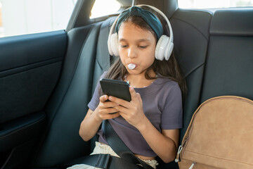 Cute schoolgirl with bubble gum scrolling in smartphone while listening to music in headphones