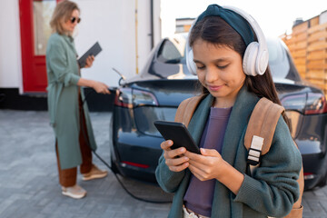 Cute girl with headphones scrolling in smartphone while waiting for her mom charging electric car