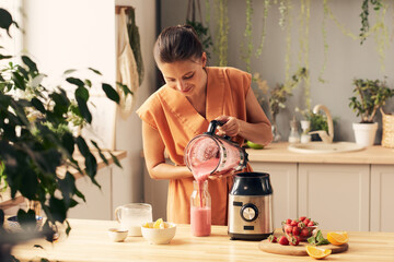 Happy woman with electric blender pouring fresh smoothie into bottle