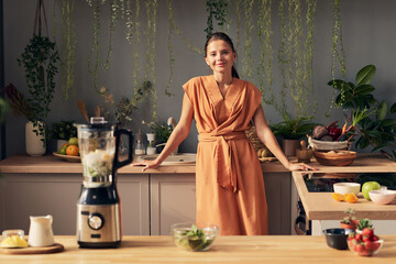Young pretty female standing in front of kitchen table with blender and ingredients for smoothie