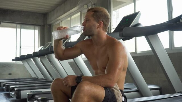 Handsome muscular man running sitting on treadmill finished exercise drinking water in fitness gym . Shirtless sport guy workout  on running Machine . bodybuilder cardio training