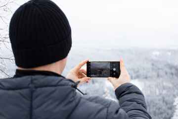 Obraz premium Close-up of a man photographing a winter landscape on the phone. Mobile photography. Winter tourism and hiking in the forest. Copy space