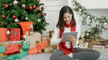 Asian woman using smart tablet sit at christmas tree xmas present gift box in cozy living room....