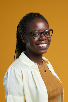 Vertical portrait of young African-American woman wearing glasses and smiling happily at camera while posing against yellow background in studio