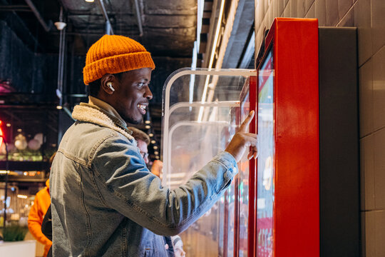 Positive African-American guy in orange knitted hat with wireless earphones orders food through self-service kiosk in cafe