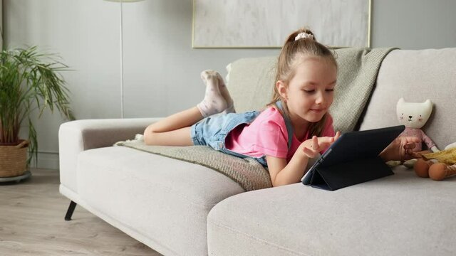 Smiling little girl lying on sofa playing online games, web surfing information, using funny applications on tablet.