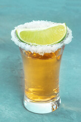 Gold tequila shot, with a salt rim and a lime slice, an angle close-up view on a blue background