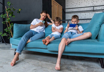 Adult woman and two children are sitting on blue sofa. Mom and son use phone and do not pay attention to girl sitting next to them.