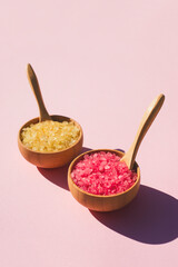 Wooden bowls with a spoons filled with pink and yellow bath sea salt. Beauty treatment for spa and wellness on pink background. Skincare natural cosmetic concept for body care
