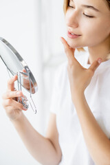 Bathroom treatment. Skin beauty. Facial procedure. Attractive satisfied woman admiring flawless smooth face in mirror on defocused light background.