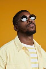 Fototapeta premium Vertical portrait of handsome African-American man wearing sunglasses while posing against yellow background in studio