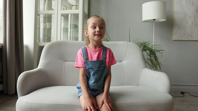 Five years little girl with pretty face smiling looking at camera at home sitting on chair near sofa, talking to webcam making online video call or recording vlog, waving hand.