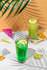 beautiful still life composition with two transparent glasses of summer drink. green lemonade with lime, mint, lemon and citrus on a tropical background with shadows of a palm tree
