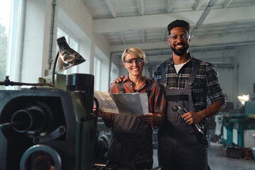 Portrait of young biracial industrial colleagues working indoors in metal workshop, smiling and looking at camera.