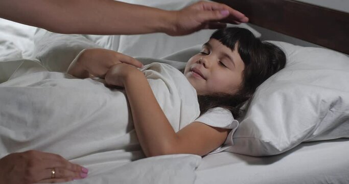 Close up  of little girl 6 years old with long brown hair falling asleep  in bed with white linens. Mom's hands are straightening her blanket.  Preschool girl  going to bed. Good night concept 4k 