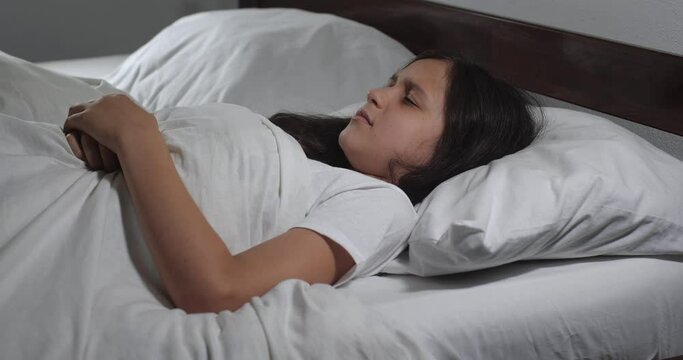 Close up   of  girl 9 years old with long brown hair  lying in bed with white linens in light room suffering from stomach-ache. Teenager suffers from pain holding stomach with hands   4K 