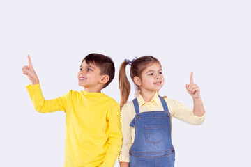 Picture of two schoolchildren boy and girl standing shoulder to shoulder with joy facial expression holding their index fingers up.