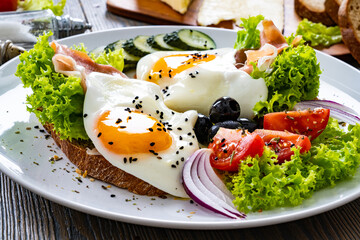 Sunny side up eggs on toasted bread and prosciutto di Parma  with fresh green vegetables, mini tomatoes and black olives on wooden table
