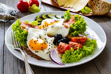 Sunny side up eggs on toasted bread and prosciutto di Parma  with fresh green vegetables, mini tomatoes and black olives on wooden table
