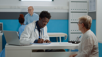 Fototapeta premium African american doctor writing checkup on notebook while consulting senior woman at desk. Black medic checking on patient healthcare while nurse and elder man talking in background
