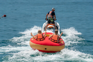 Happy people are going to swim on an air mattress behind a jet ski. Tourists ride the inflatable watercraft boat. Sea attraction. Jet ski skating people on a large Inflatable raft. Aerial copter view.