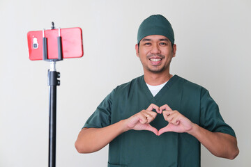Male hospital nurse giving love heart sign in front of mobile phone. Online consultation concept