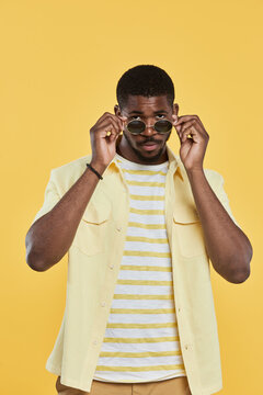 Vertical portrait of trendy African-American man wearing sunglasses and looking at camera while standing against yellow background in studio