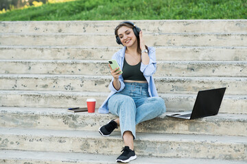 Happy and attractive caucasian girl sitting with a laptop on the steps and holding a phone, listening to music on headphones