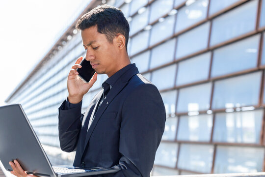 Businessman with laptop talking on smart phone outside office building during sunny day
