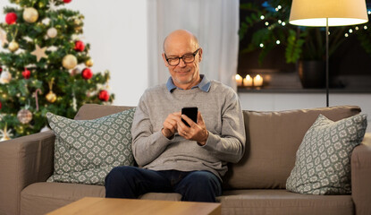 Fototapeta premium technology, people and winter holidays concept - happy smiling bald senior man texting on smartphone at home in evening over christmas tree on background