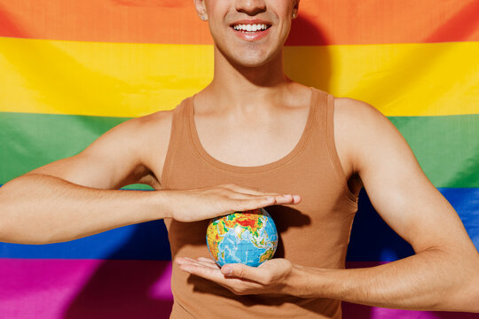 Cropped young queer smiling satisfied fun blond latin gay man 20s wear beige tank shirt hold in hands Earth world globe stand on rainbow flag background studio portrait People lgbt lifestyle concept.