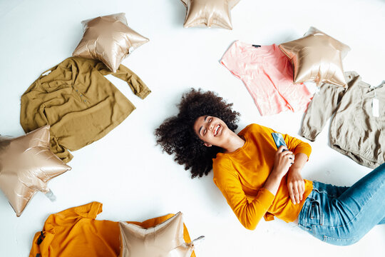 Cheerful woman with mobile phone amidst clothes and foil balloons on floor