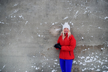 Active senior woman runner standing against concrete wall outdoors in snowy winter, looking at camera.