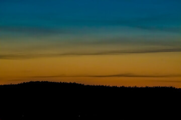 Natural ombre of evening sky, Vilnius, Lithuania. Blue and orange colors, small dark clouds, forest in the shadows. Selective focus on the pattern, blurred background.