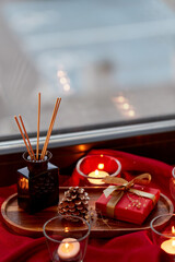 holidays, decoration and celebration concept - christmas gift, cup of coffee, candles and aroma reed diffuser on red tablecloth on window sill at home