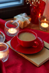 holidays, decoration and celebration concept - cup of coffee in red ceramic cup with saucer, burning candles and christmas gift on window sill at home