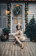 Young beautiful woman wearing knitted hat and white sweater sitting on porch of holiday decorated...