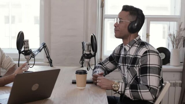 Young men talking and smiling while sitting at table while radio broadcasting in studio epspb. Closeup view of two American people look with smile and talk, record business podcast and sit at desk in