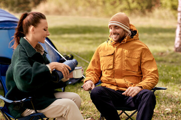 Fototapeta premium camping, tourism and travel concept - happy couple with thermos pouring tea to cup at tent camp